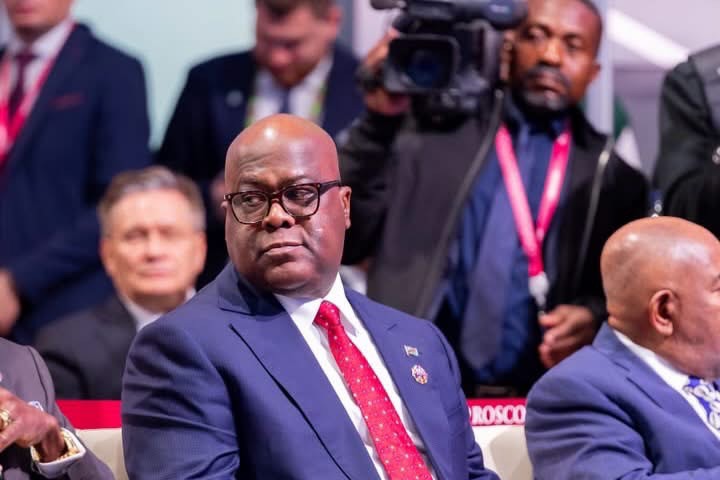 Félix Tshisekedi assis parmi les participants à une rencontre, avec des journalistes à l’arrière-plan.