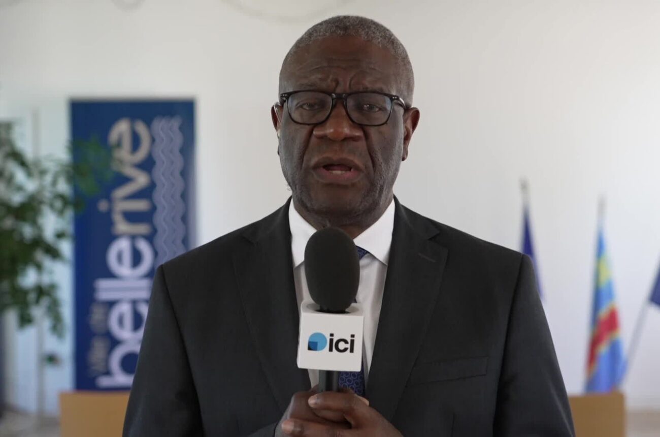 Denis Mukwege, en costume sombre et lunettes, répond à une interview au micro d’ICI, devant les drapeaux de la France, de la RDC et de l’Union européenne.