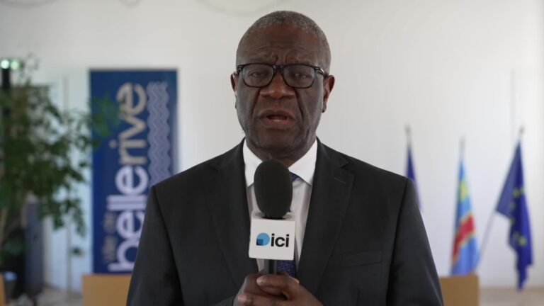 Denis Mukwege, en costume sombre et lunettes, répond à une interview au micro d’ICI, devant les drapeaux de la France, de la RDC et de l’Union européenne.